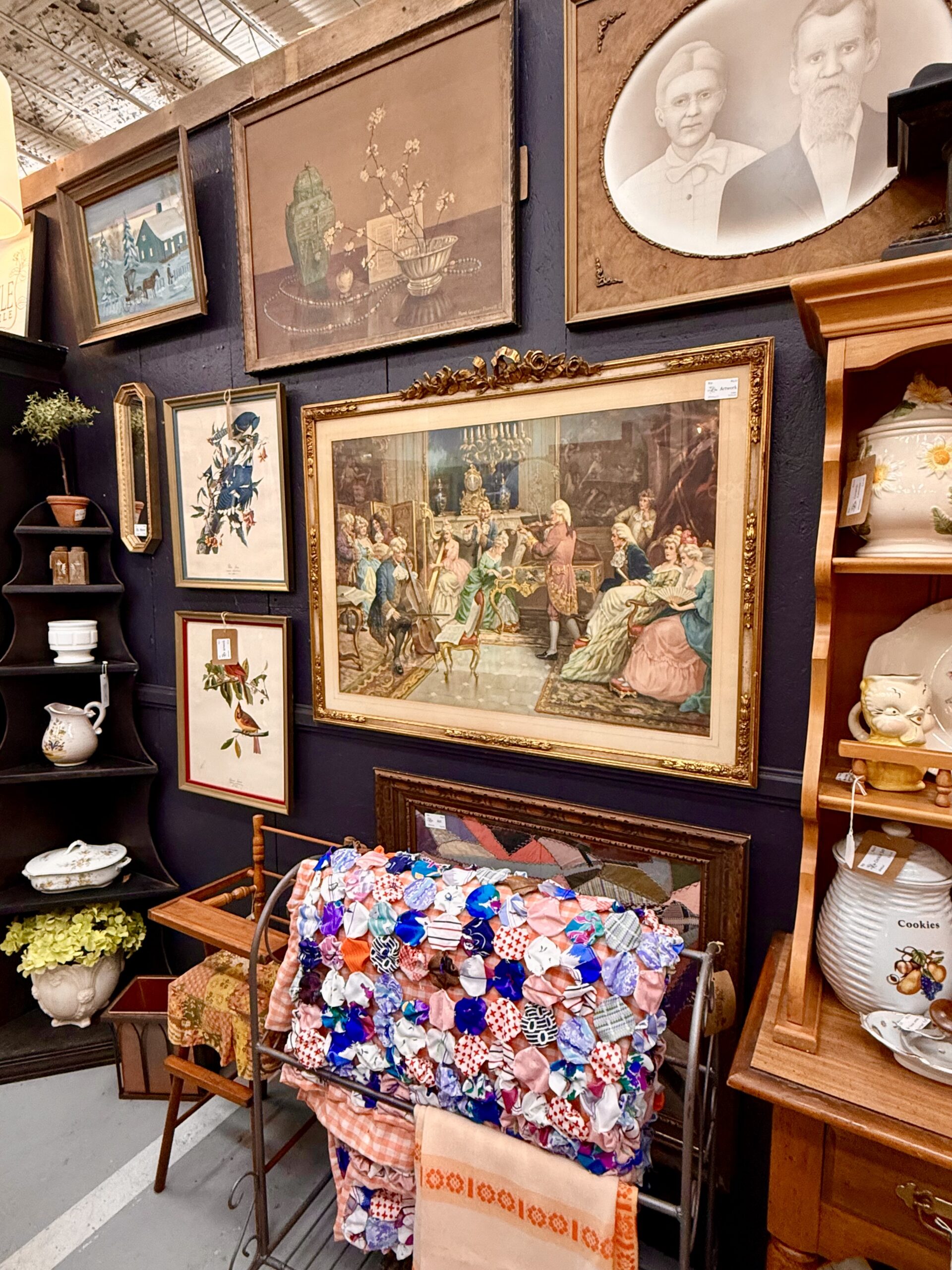 Gallery wall of vintage framed art above antique furniture with patchwork quilt and decorative shelves in the booth.